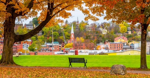 A small town covers a hillside with brick buildings and trees covered in fall foliage. In the image foreground, a bench and fall foliage frame the view.