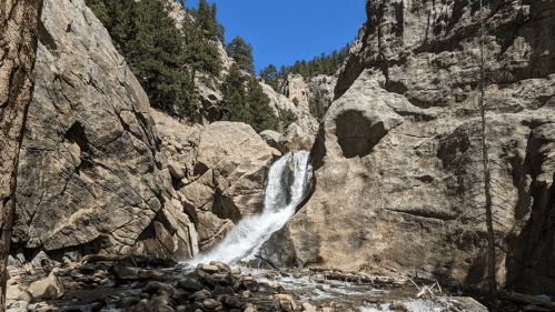 A waterfall cascades over rocky cliffs, surrounded by trees under a clear blue sky.