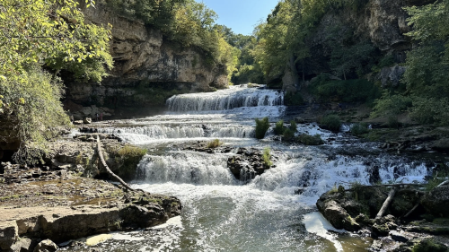 A serene waterfall cascades over rocky ledges, surrounded by lush greenery and a clear blue sky.