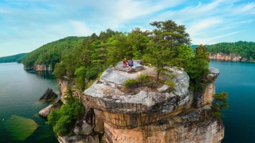 Aerial view of a rocky cliff surrounded by water and lush greenery, with two people sitting on top.
