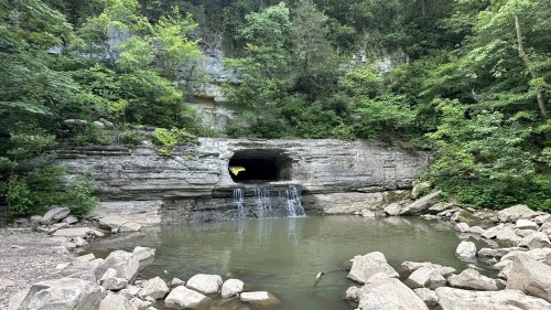 A rocky landscape with a small waterfall flowing into a calm pool, surrounded by lush green trees and cliffs.
