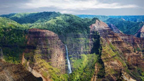 A stunning view of lush green mountains with a waterfall cascading down rocky cliffs under a blue sky.