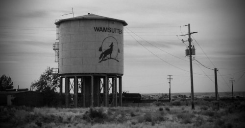 A black-and-white photo of a large water tower in the middle of the desert. The water tower says "Wamsutter" with an image of a wolf howling to the moon.