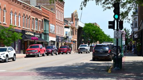 A busy street lined with shops and parked cars, featuring traffic lights and green trees on a sunny day.