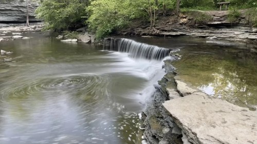 A serene waterfall cascades into a calm pool, surrounded by lush greenery and rocky terrain.