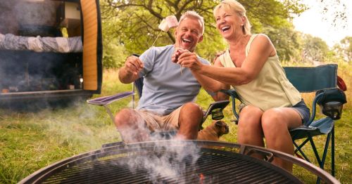 A retired couple in Indiana, roasting marshmallows over a campfire, with a camper van and trees in the background.