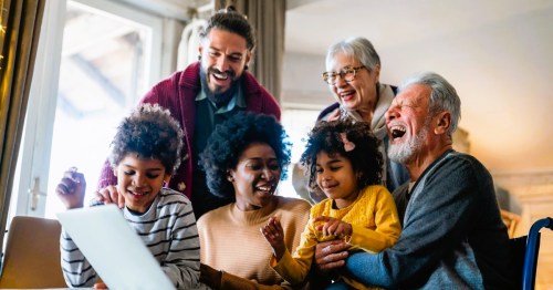 A joyful family with two children, their mother, a grandfather, and two elderly women, gathering around a laptop, sharing laughter and smiles.