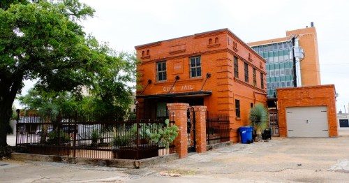 Historic city jail building in a small town in Texas, made of red brick, surrounded by a fence and greenery.