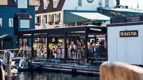 A waterfront restaurant with outdoor seating, bustling with people, surrounded by boats and buildings under a clear blue sky.