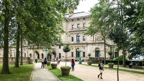 A historic building surrounded by trees, with people walking along a path in a park-like setting.