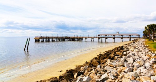 A tranquil beach with views of the crystal-clear waters at St. Simons Island in Georgia, featuring a wooden pier and rocky shoreline.
