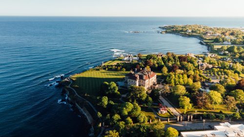 Aerial view of a coastal landscape featuring a large mansion surrounded by lush greenery and the ocean in the background.