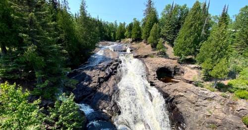 Cross River Falls cascades heavily down jagged, dark rocky ledges surrounded by lush green trees in Schroeder, MN.