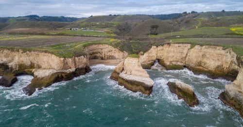 A dark turquoise ocean meets tall, sandy cliffs with hills of greenery and a long highway beyond.
