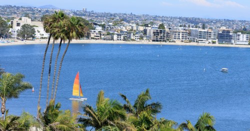 A distant sailboat in calm blue water on a sunny summer afternoon. A SoCal city is visible in the background.