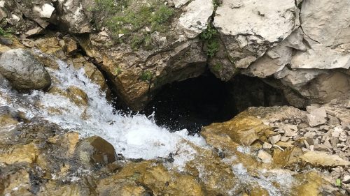 A rocky landscape with a small waterfall flowing into a dark cave entrance surrounded by stones and greenery.