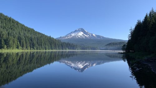 A serene lake reflects a snow-capped mountain surrounded by lush green forests under a clear blue sky.
