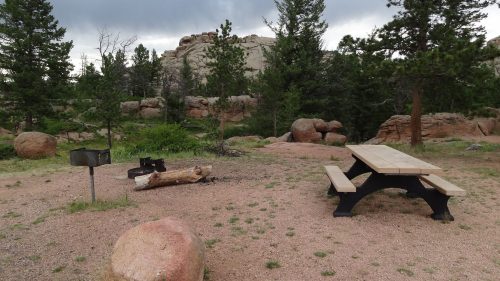 A picnic area with a wooden table, fire pit, and rocky landscape surrounded by trees under a cloudy sky.