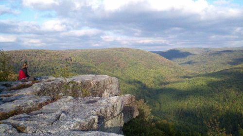 A person sits on a rocky ledge overlooking a lush green valley and rolling hills under a partly cloudy sky.