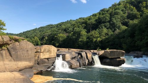A serene river scene with cascading waterfalls and lush green hills in the background under a clear blue sky.