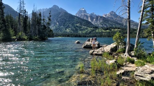 A serene lake surrounded by mountains, with two people standing on a rock near the water's edge under a clear blue sky.