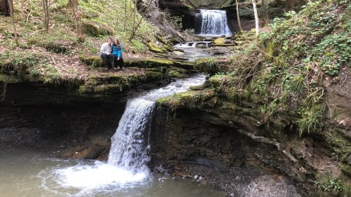 Two people sit by a small waterfall in a lush, green forest with another waterfall in the background.