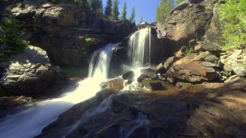 A serene waterfall cascades over rocky terrain, surrounded by lush green trees under a clear blue sky.