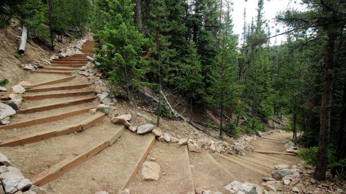 Winding wooden steps lead through a forested area, surrounded by trees and rocky terrain.