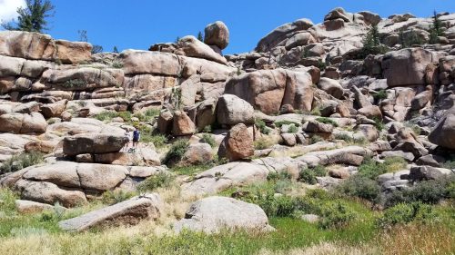 A rocky landscape with large boulders and patches of grass under a clear blue sky. A person stands on the rocks.