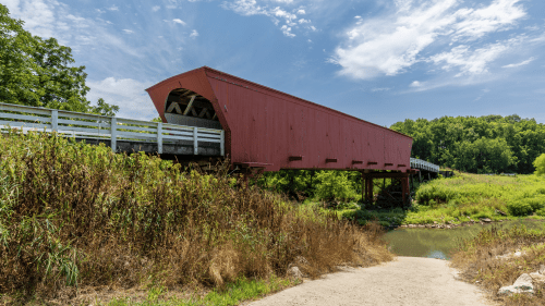 A red covered bridge spans a small creek, surrounded by lush greenery and a clear blue sky.