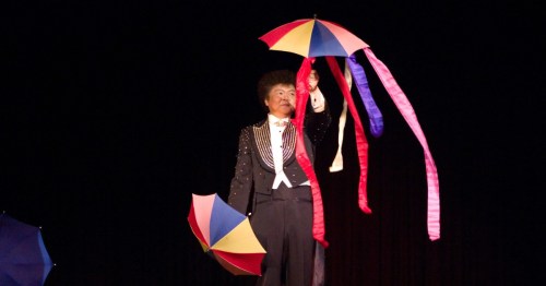 A magician in a tuxedo performs on a dark stage, holding a colorful umbrella with long, flowing ribbons attached.