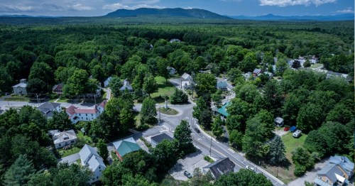 A small town in the middle of a vividly green forest is in the foreground as a few tall mountains are seen in the background.