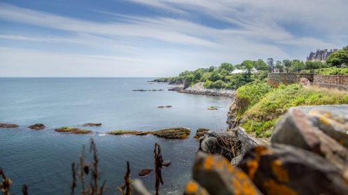 A serene coastal view with rocky shores, calm water, and lush greenery under a blue sky with wispy clouds.