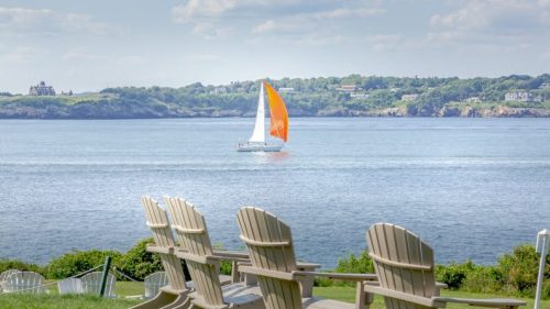 A sailboat with an orange sail glides across calm waters, with Adirondack chairs in the foreground and lush greenery in the background.