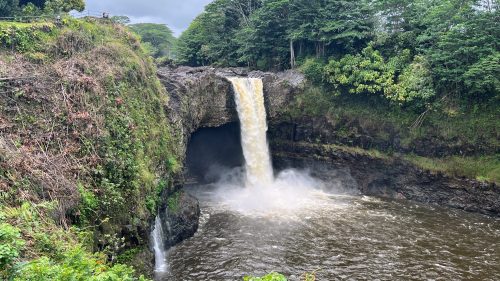 A waterfall cascades into a river, surrounded by lush greenery and rocky cliffs under a cloudy sky.