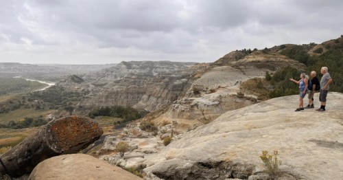 Three people stand on a rocky overlook in Theodore Roosevelt National Park viewing the vast rugged canyon.