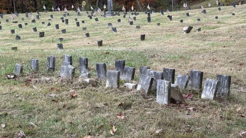 A historic cemetery at The Ridges in Athens, Ohio, where patients from the former mental hospital (1874–1993) were laid to rest. The grassy field contains numerous small, weathered gravestones, some leaning or partially sunken. American flags are placed near several markers, and autumn foliage provides a colorful backdrop.
