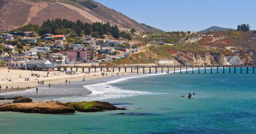 Turquoise waves roll toward Harford Pier in Avila Beach, California, with surfers nearby.