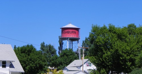 The red Washburn water tower stands tall above green trees and neighborhood homes under a bright blue sky in ND.