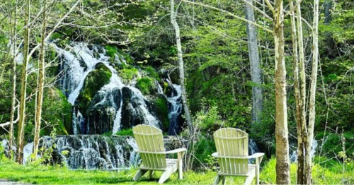 Two chairs sit just beside a rushing waterfall and flowing creek with green grass surrounding.