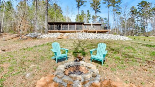 A serene outdoor scene featuring two teal chairs by a fire pit, with a house and trees in the background.