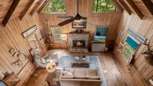 Cozy wooden living room with a stone fireplace, large windows, and rustic decor, surrounded by greenery.