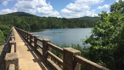 A scenic view of a bridge over a calm lake, surrounded by lush greenery and mountains under a partly cloudy sky.