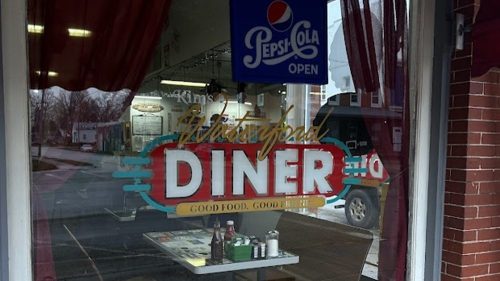A diner window with a "Pepsi Cola Open" sign and a wooden bench outside, showcasing a welcoming atmosphere.