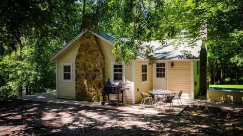 A cozy yellow cabin with a stone chimney, surrounded by trees, featuring a patio with a table and chairs.