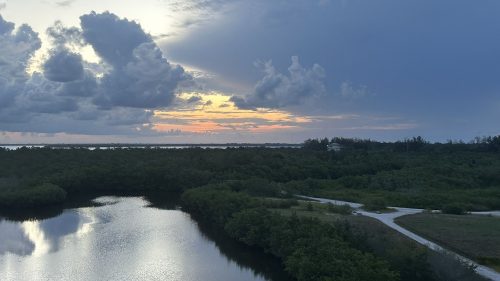 A serene landscape featuring a calm waterway, lush greenery, and a dramatic sky at sunset with clouds.