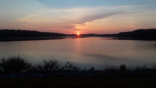 A serene sunset over a calm lake, with silhouettes of trees and gentle ripples on the water's surface.