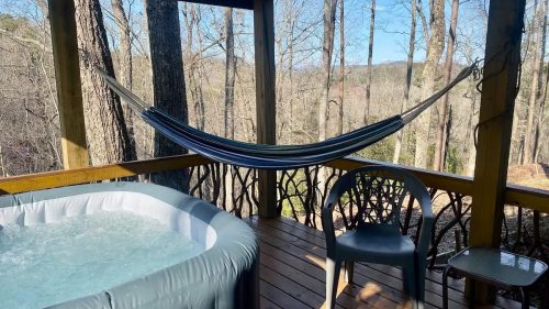 A cozy deck with a hot tub, hammock, and chairs, surrounded by trees and a scenic view.