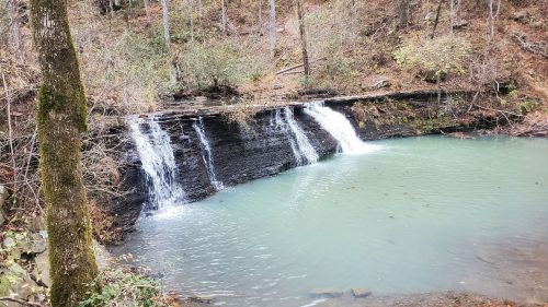 A serene waterfall cascades into a calm, turquoise pool surrounded by autumn foliage and trees.