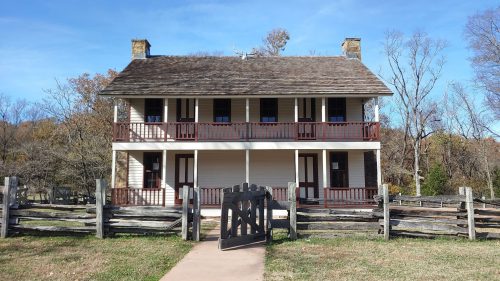 Historic two-story house with a wooden fence and gate, surrounded by trees and a clear blue sky.